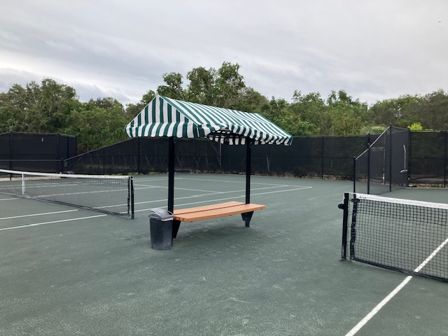 A striped canvas canopy bench beside a tennis court, the fabric faded and sagging over a plain wood plank seat.